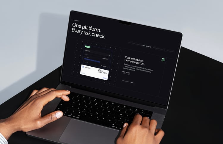 A person using a laptop displaying a dark-themed Baselayer interface with the headline “One platform. Every risk check.” and a panel showing connected data and a risk score.