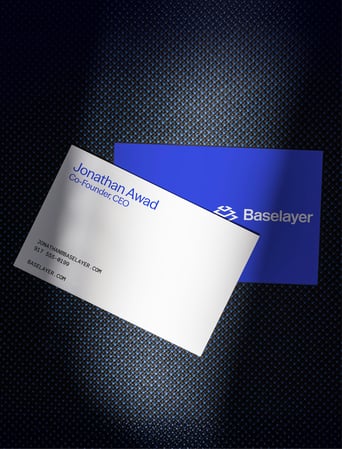 Two Baselayer business cards on a textured dark surface, one showing Jonathan Awad’s contact details and the other displaying the Baselayer logo on a blue background.
