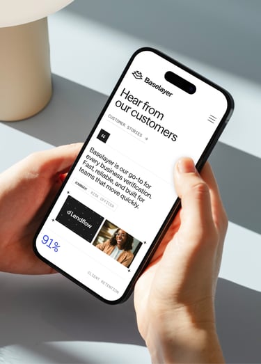A person holding a smartphone displaying a Baselayer webpage with customer testimonials, including a quote, customer details and featured customer logos and images.